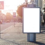 Blank advertisement on tiled walkway with road signs and building in town on sunny day - Photo by Julia Filirovska on Pexels