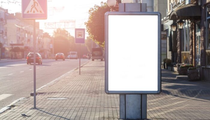 Blank advertisement on tiled walkway with road signs and building in town on sunny day - Photo by Julia Filirovska on Pexels